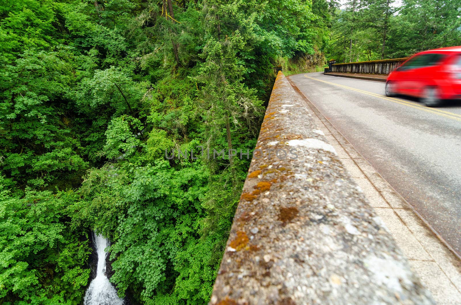 Waterfall in a lush green forest in Oregon next to historic Highway 30 with a red blurry car