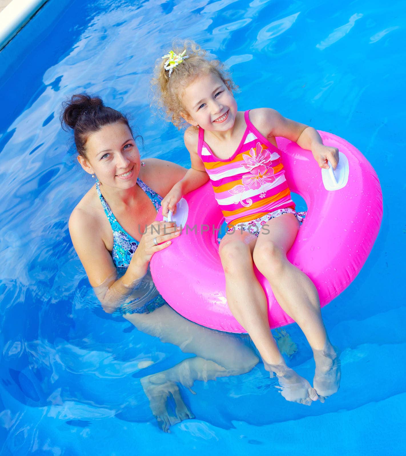 Pretty little girl in an pink life preserver with her mother in swimming pool outdoors. Vertical view.