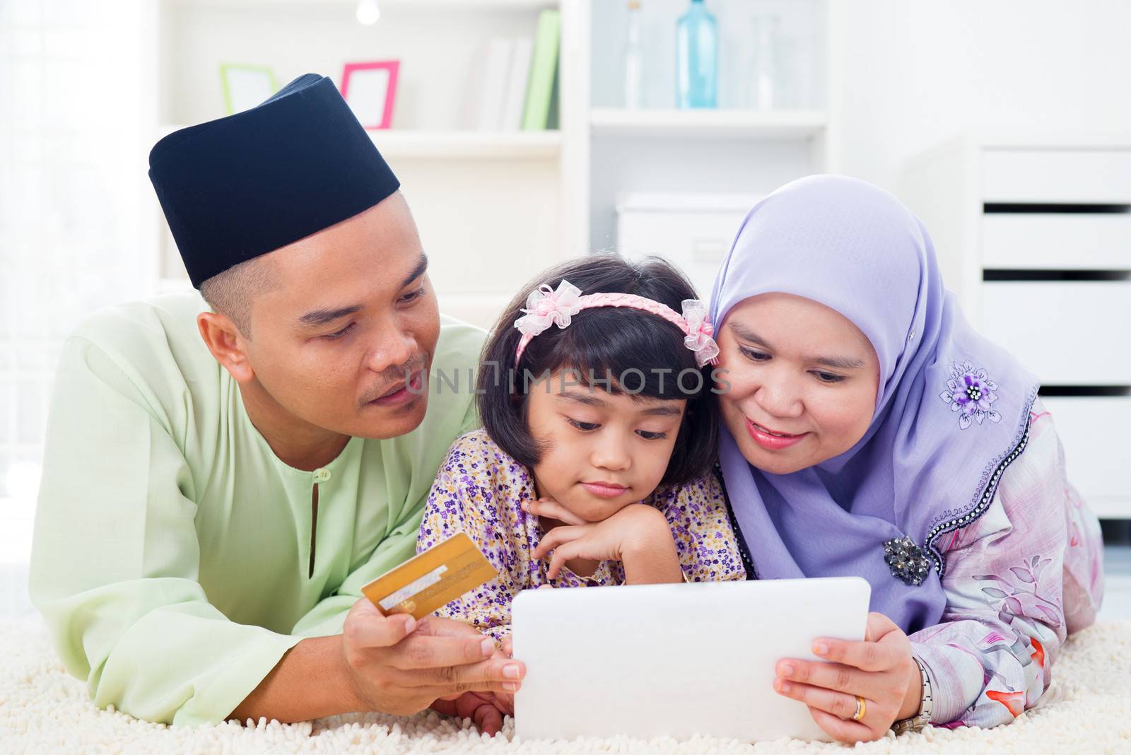 Southeast Asian family using tablet pc computer online shopping with credit card at home. Muslim family living lifestyle. Happy smiling Malay parents and child.