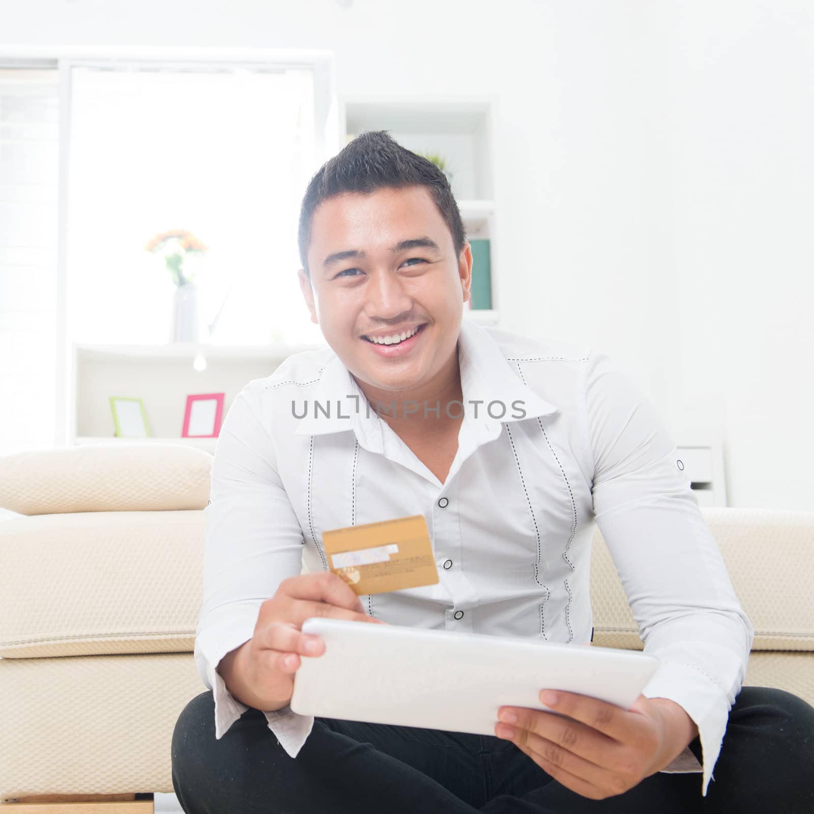 Attractive Southeast Asian man using a credit card to shop on the internet with a tablet computer. Asian male model at home.