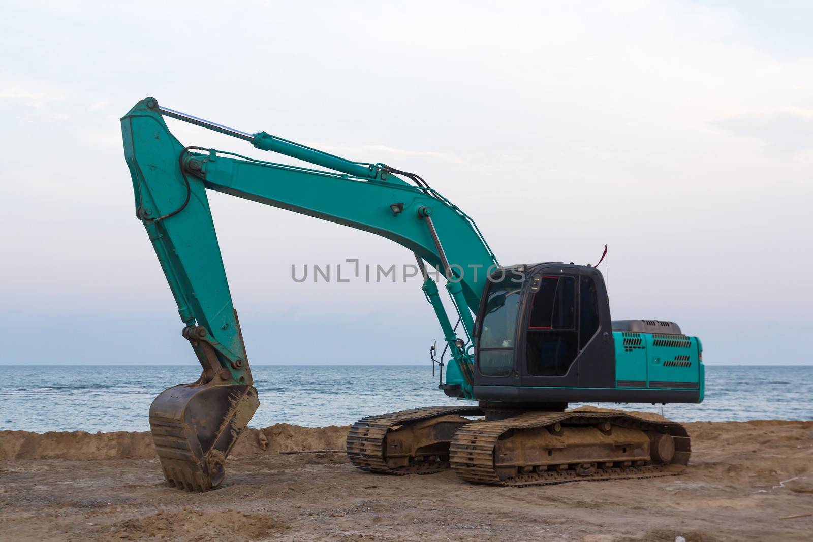 Excavator standing in sandpit with raised bucket at seaside