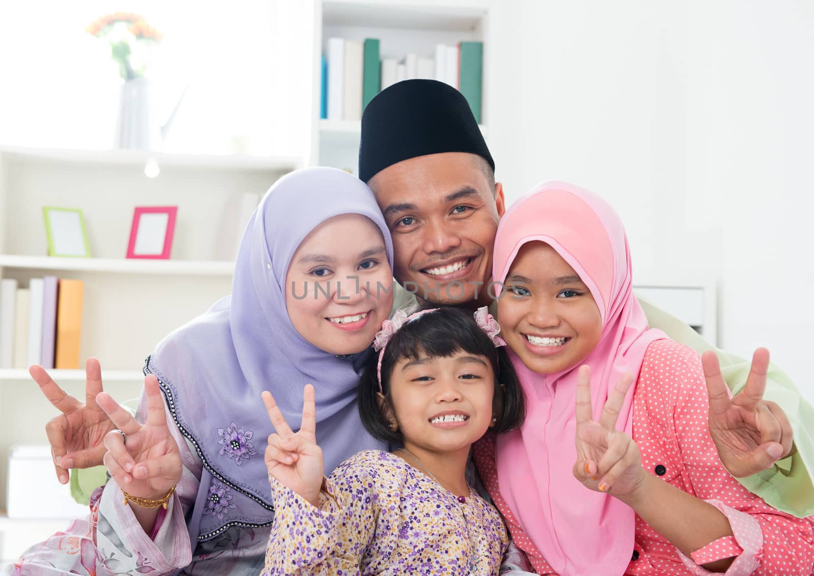 Happy Asian family at home. Muslim family showing v victory hand sign and having fun. Southeast Asian parents and children smiling.