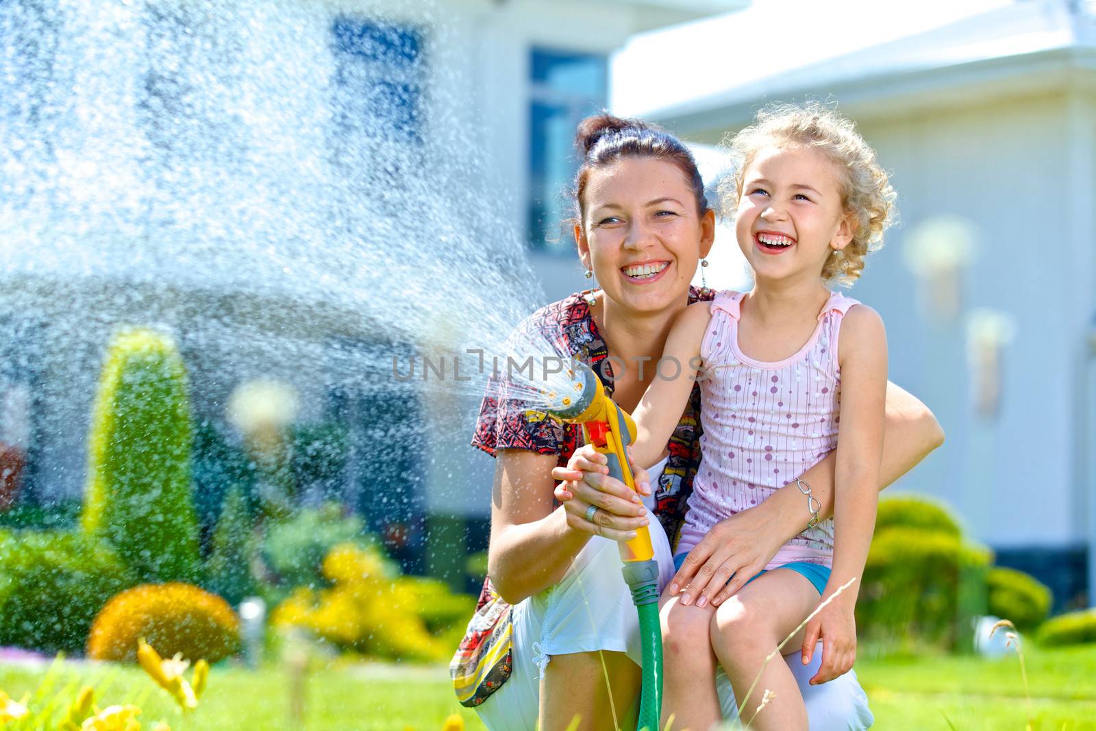 Portrait of little gardener girl with mother watering flowers on the lawn near cottage.