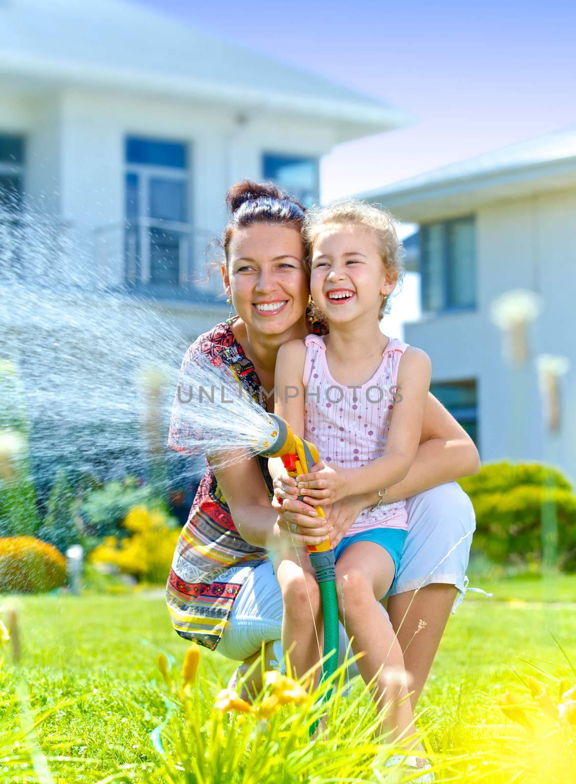 Little gardener girl with mother watering flowers on the lawn near cottage. Vertical view