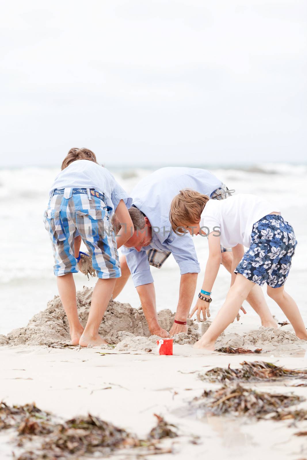 father and sons on the beach playing in the sand by juniart