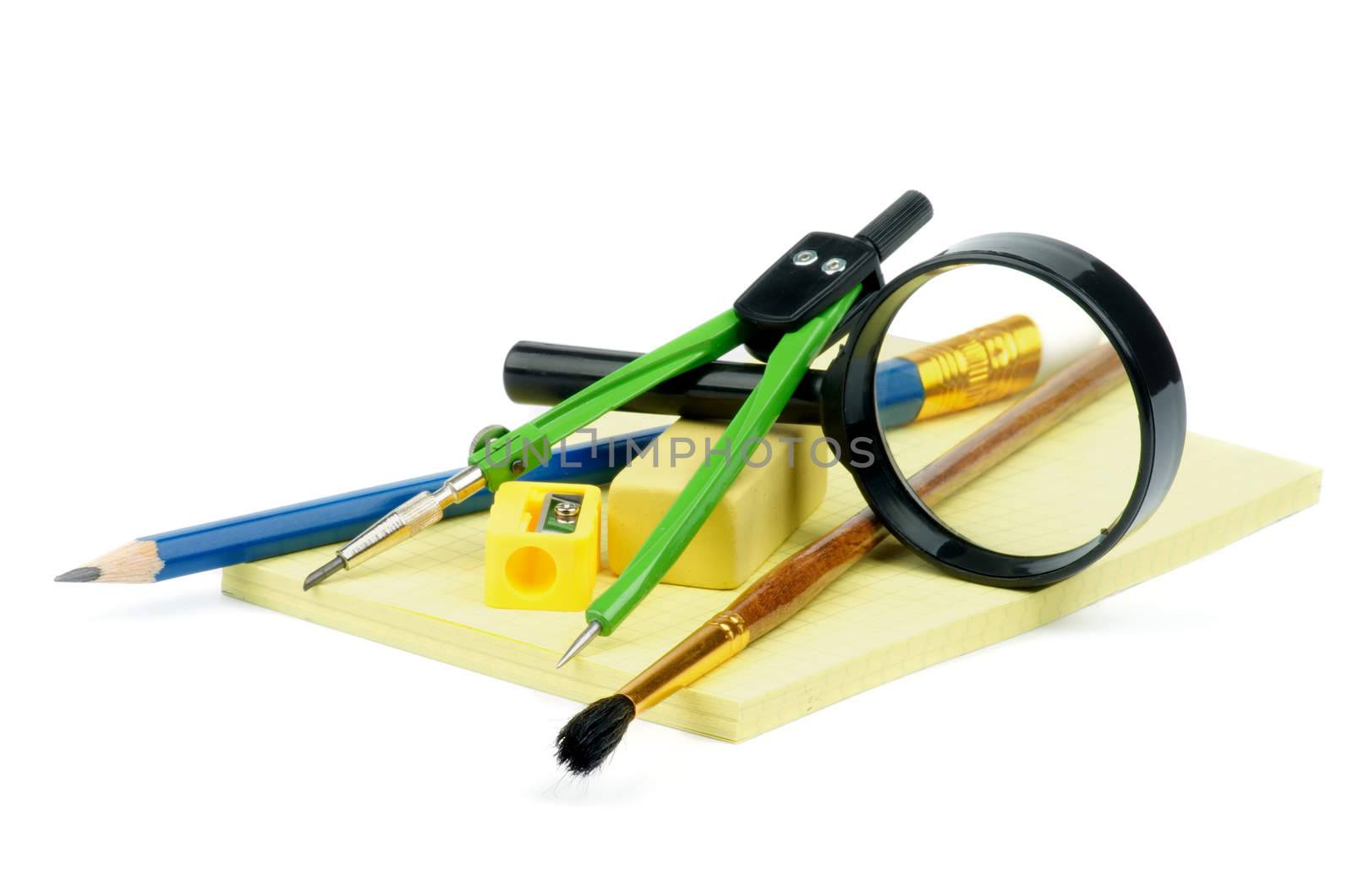 Arrangement of Pencil, Pen, Paintbrush, Pencil Sharpener, Eraser, Drawing Compass and Magnifier on Yellow Note Pad isolated on white background