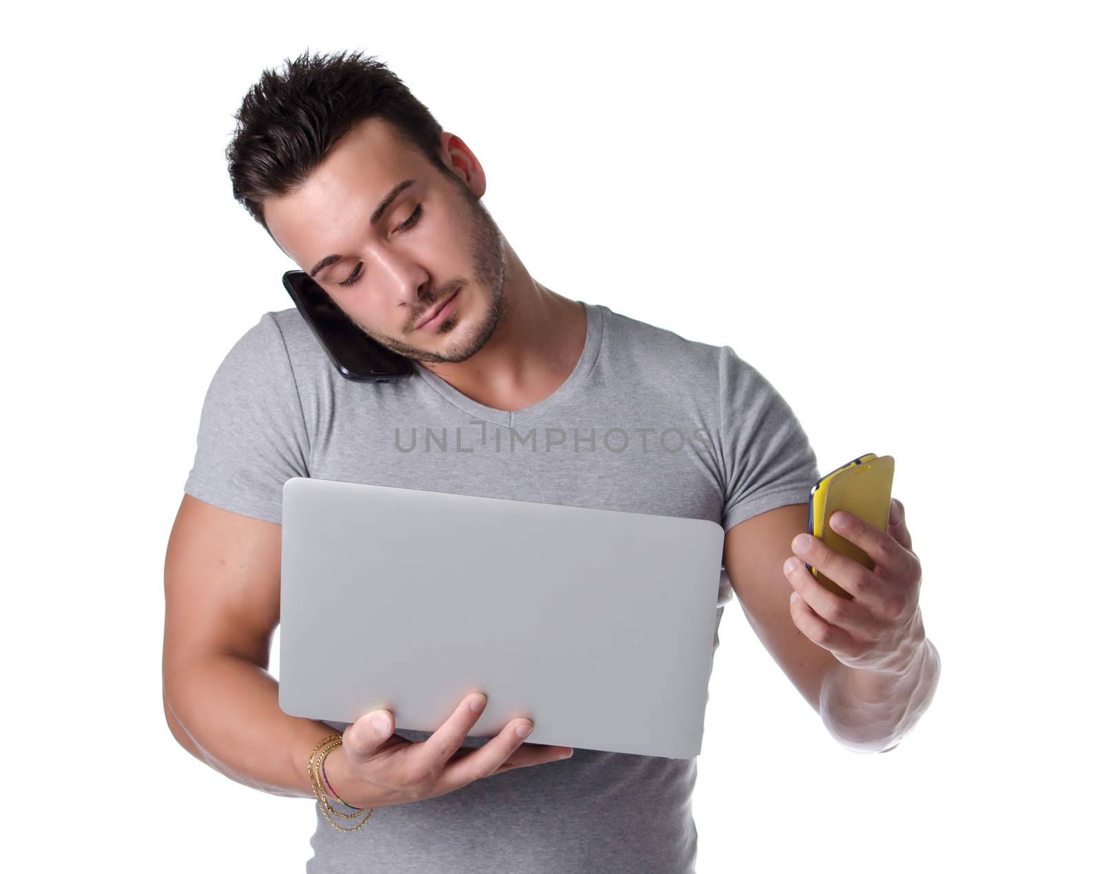 Too much technology. Young man using two mobile phones and one laptop computer, isolated on white