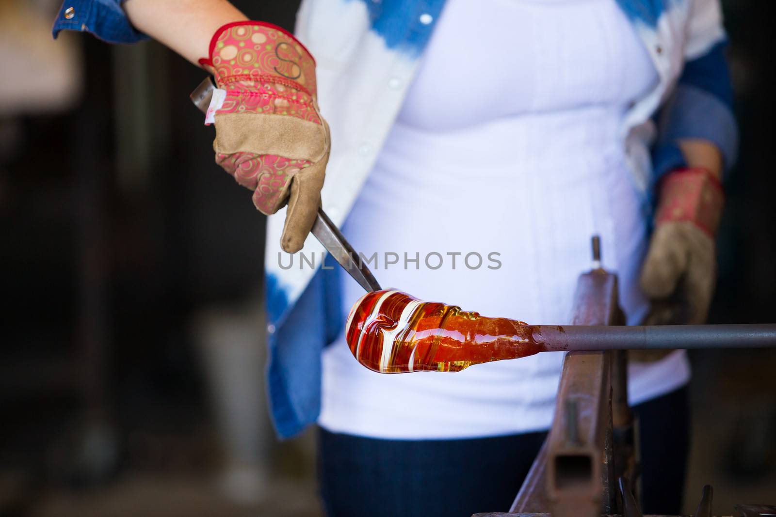 A female is blowing glass and using a wooden shaping tool to form the shape she wants the glass to end up as. The glass is hot and molten so she has to use water with the wood.