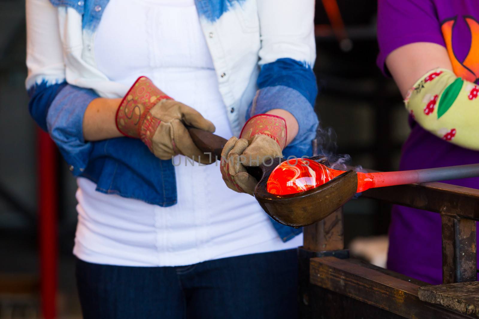 A female is blowing glass and using a wooden shaping tool to form the shape she wants the glass to end up as. The glass is hot and molten so she has to use water with the wood.