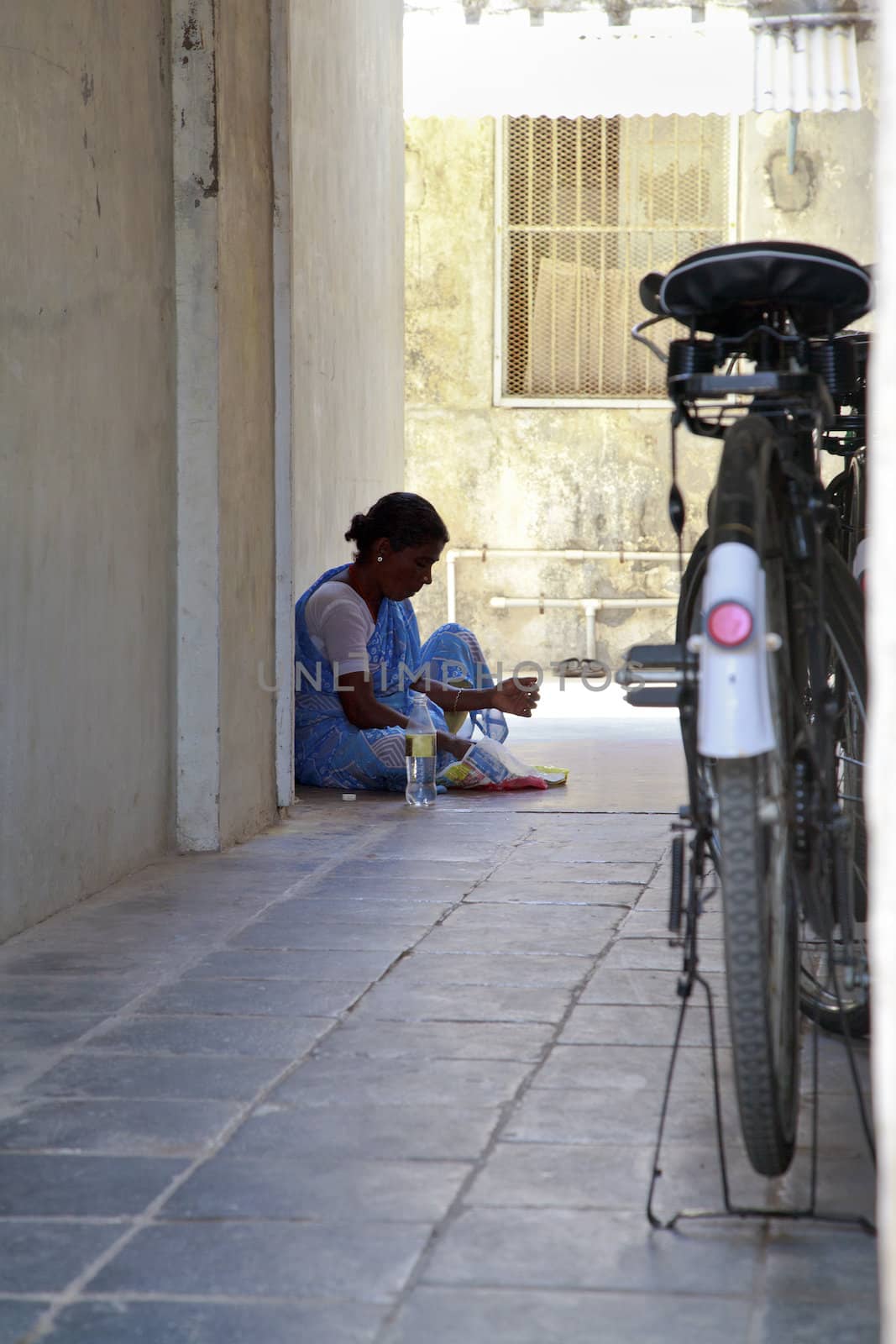 Vertical capture of an Indian maid sat on the floor in the shade inside an alleyway having lunch which has been wrapped in newsprint. Typical shot in Indian with bicycle parked close by