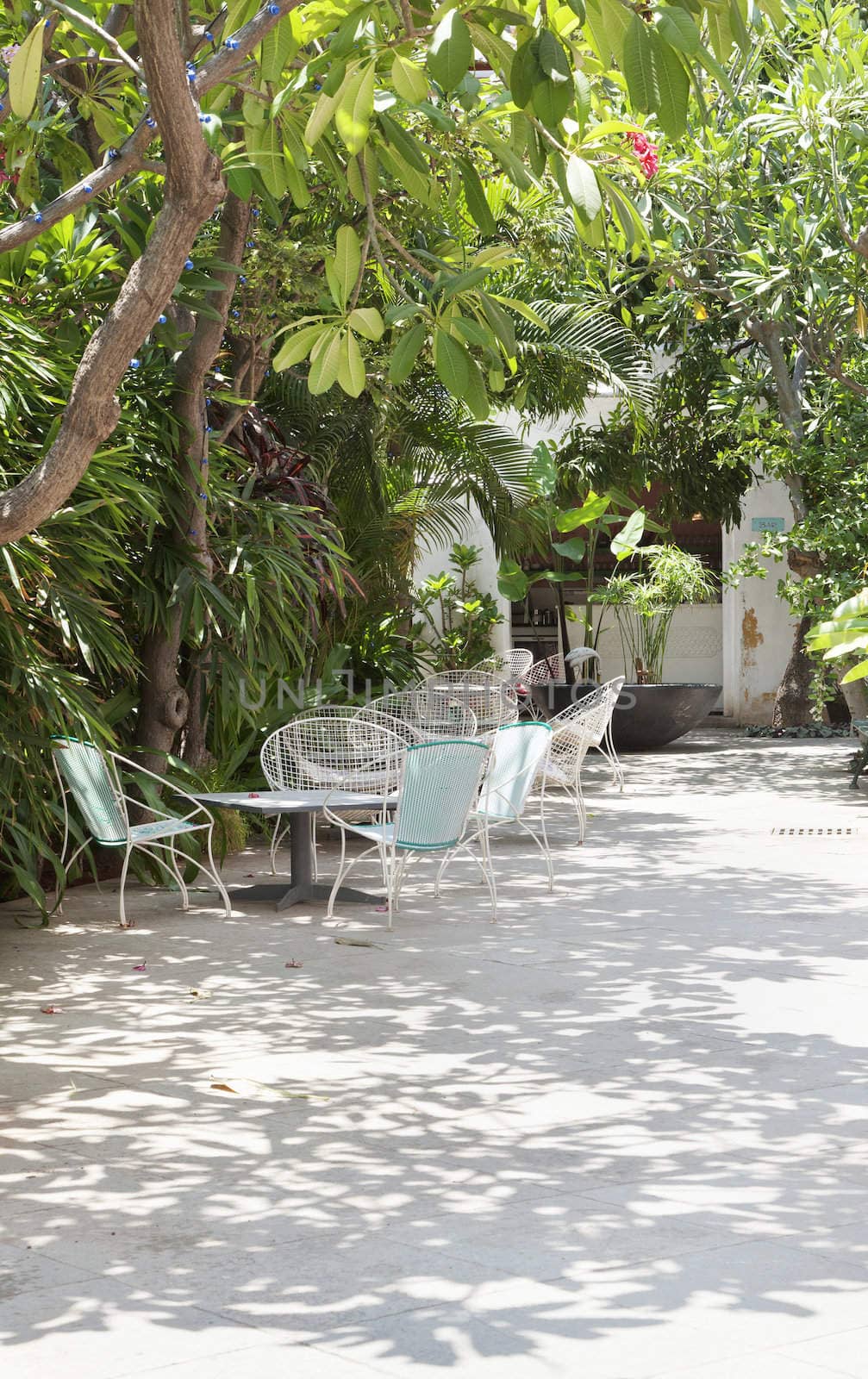 vertical image of shaded patio area with hard standing, tables and wire bucket garden chairs. Mature tree lined hedges and out buildings in distance