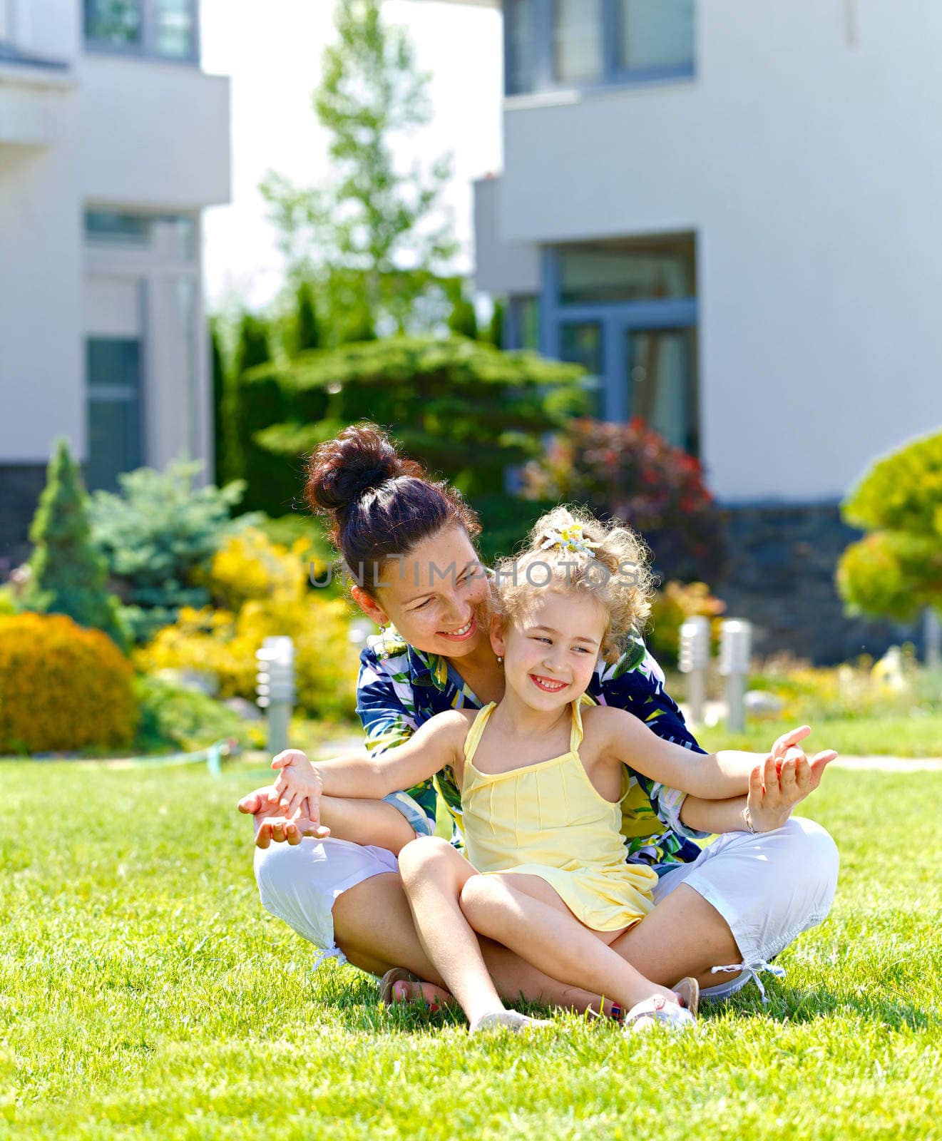 Happy girl with her mother sitting on the lawn in front new cottage. Vertical view