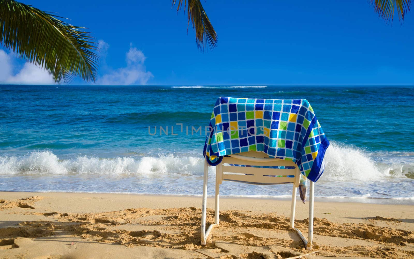 White beach chair under palm leaves by the ocean, with towel