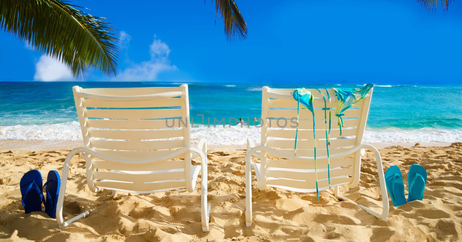 Two white beach chairs under palm leaves by the ocean, with bikini and flip flops and couple in the ocean on background