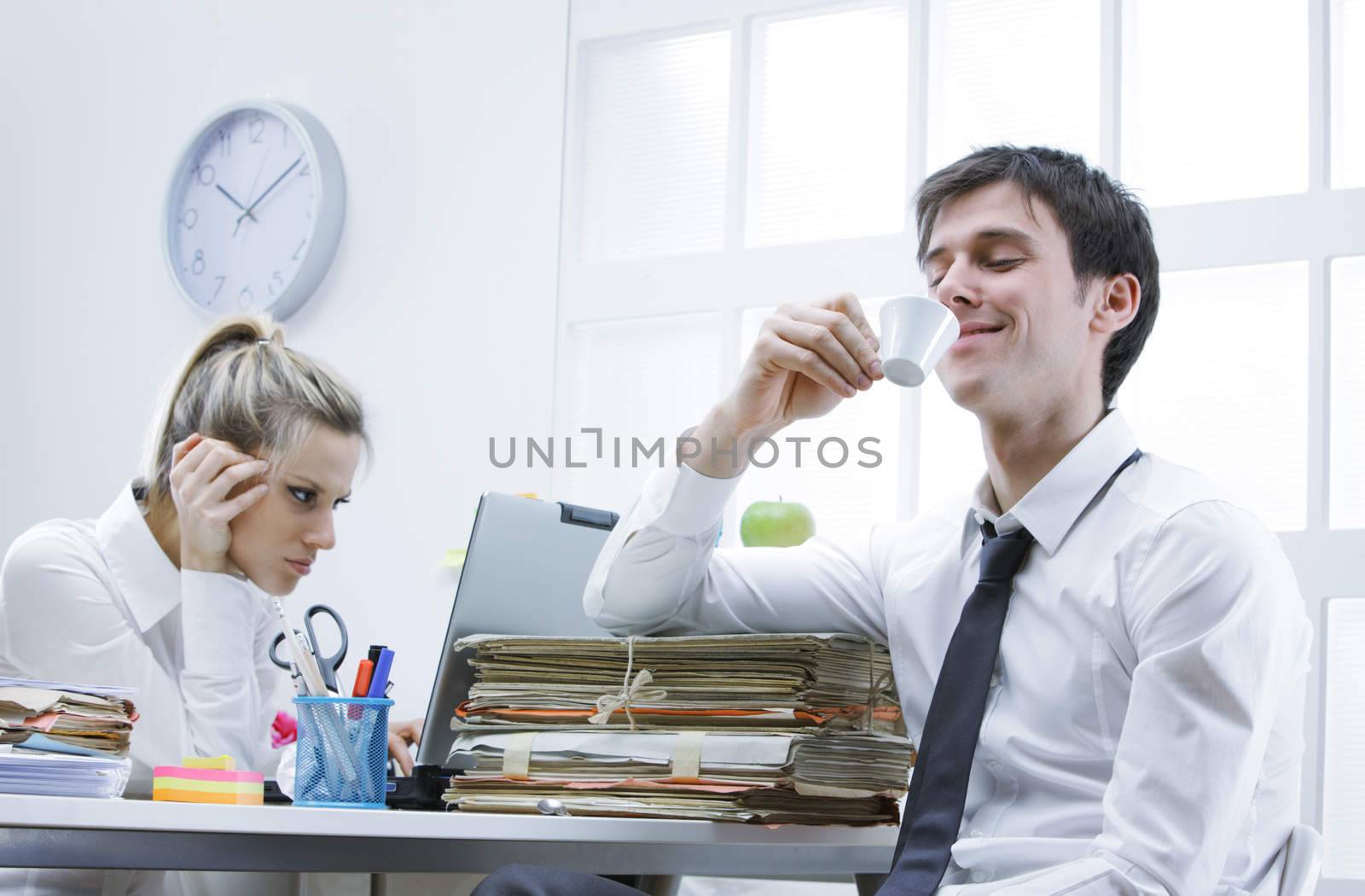 Business man enjoys a hot cup of coffee in the office, annoyed female colleague in the background
