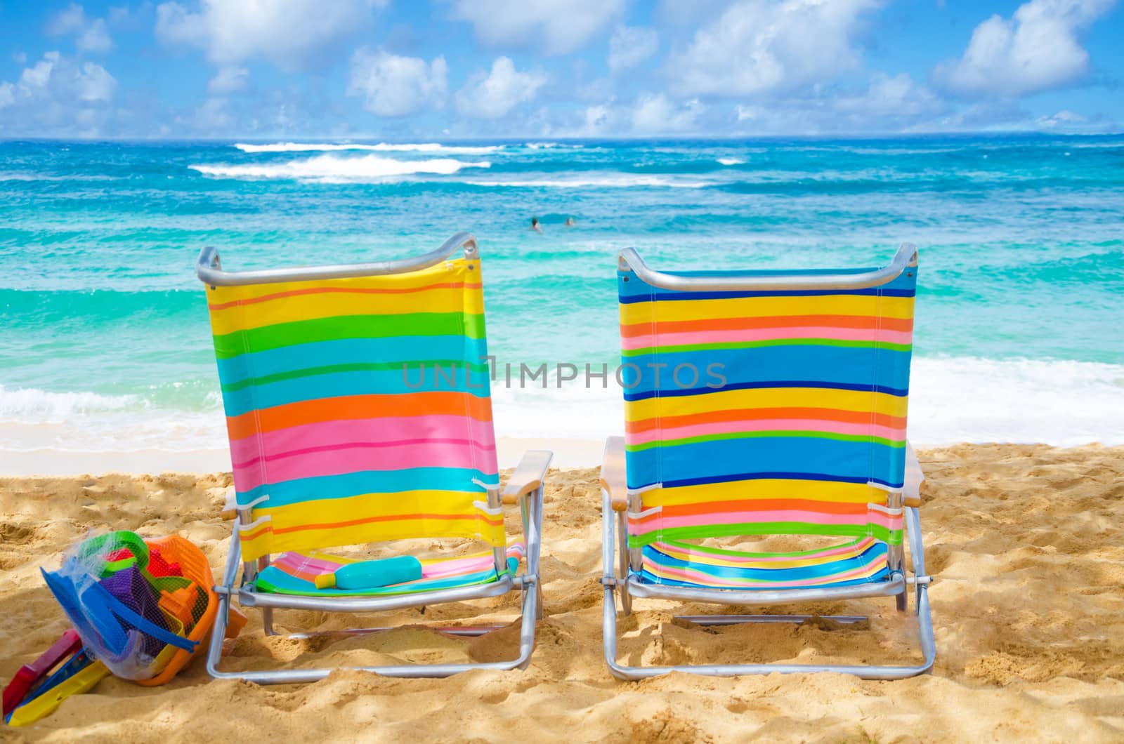 Two colorful beach chairs under by the ocean with sunscreen and kid's beach toys, with couple in the ocean on background