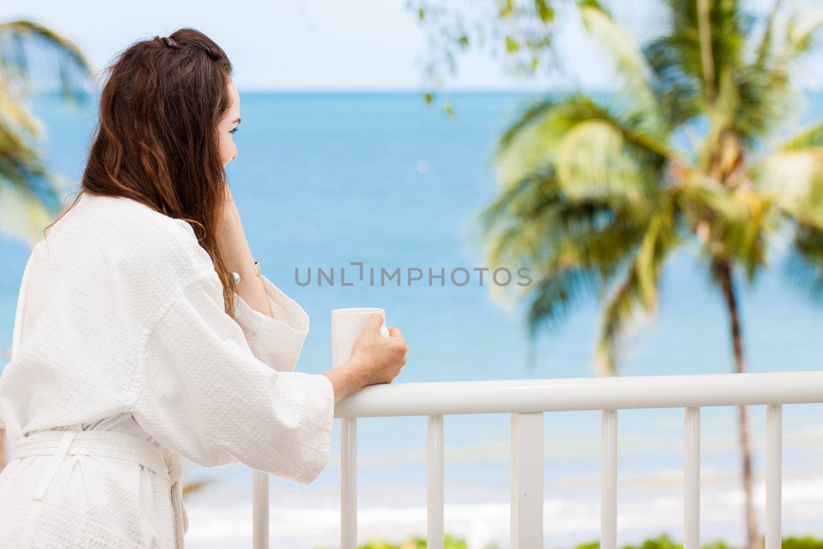 Woman in bath robe looking at beautiful ocean view  from a tropical balcony with morning coffee.