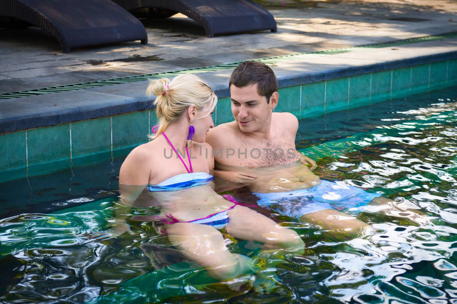 Happy smiling couple looking on each other while relaxing on the edge of a outdoor swimming pool 