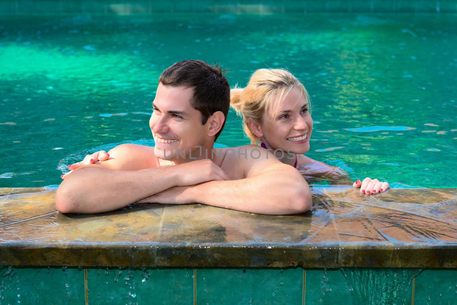 Happy smiling couple looking aside while relaxing on the edge of a outdoor swimming pool 