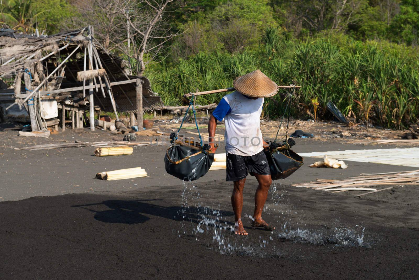 Manual male worker spreads sea water on black volcanic sand for salt production in Amuk Bay, Bali, Indonesia. It is a unique tradition dating back over 900 years. 