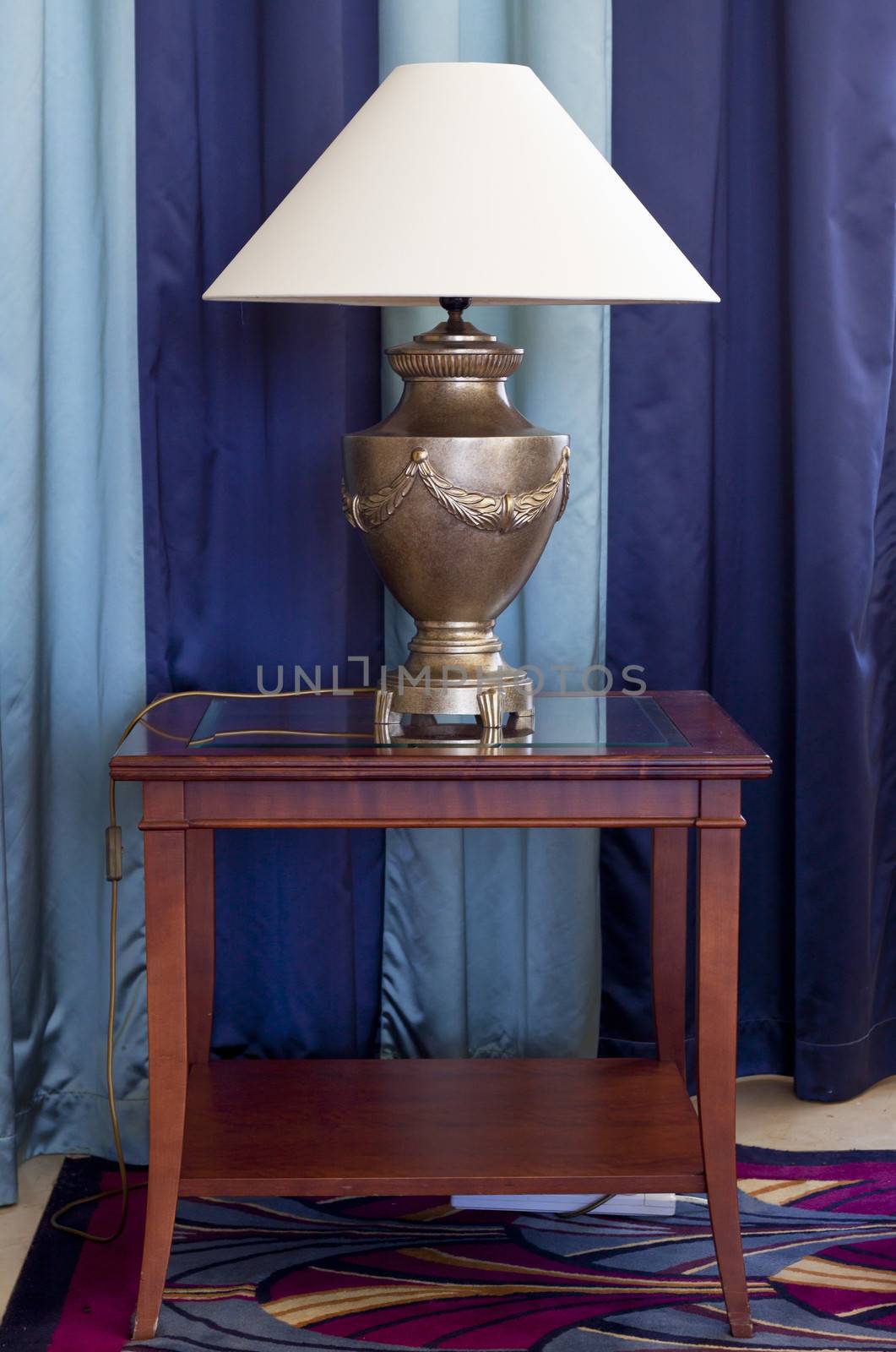 A living room lamp on a side table placed on a carpet and in front of a window with striped curtains
