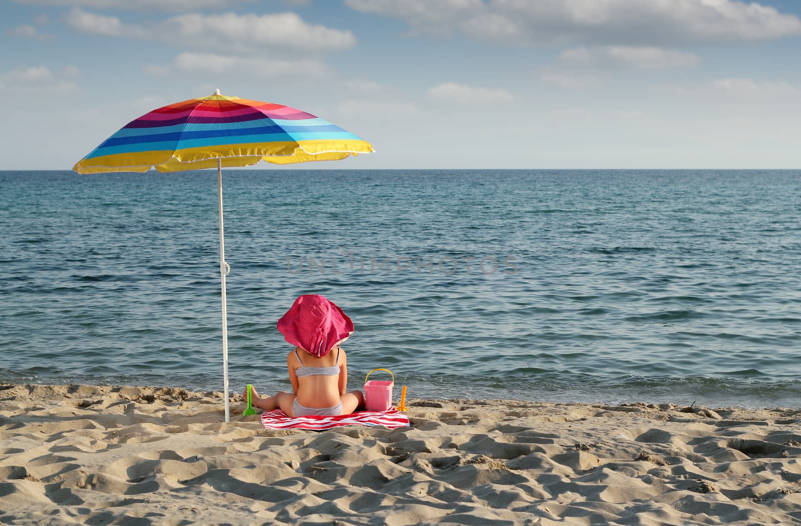 little girl with hat sitting under sunshade on beach by goce