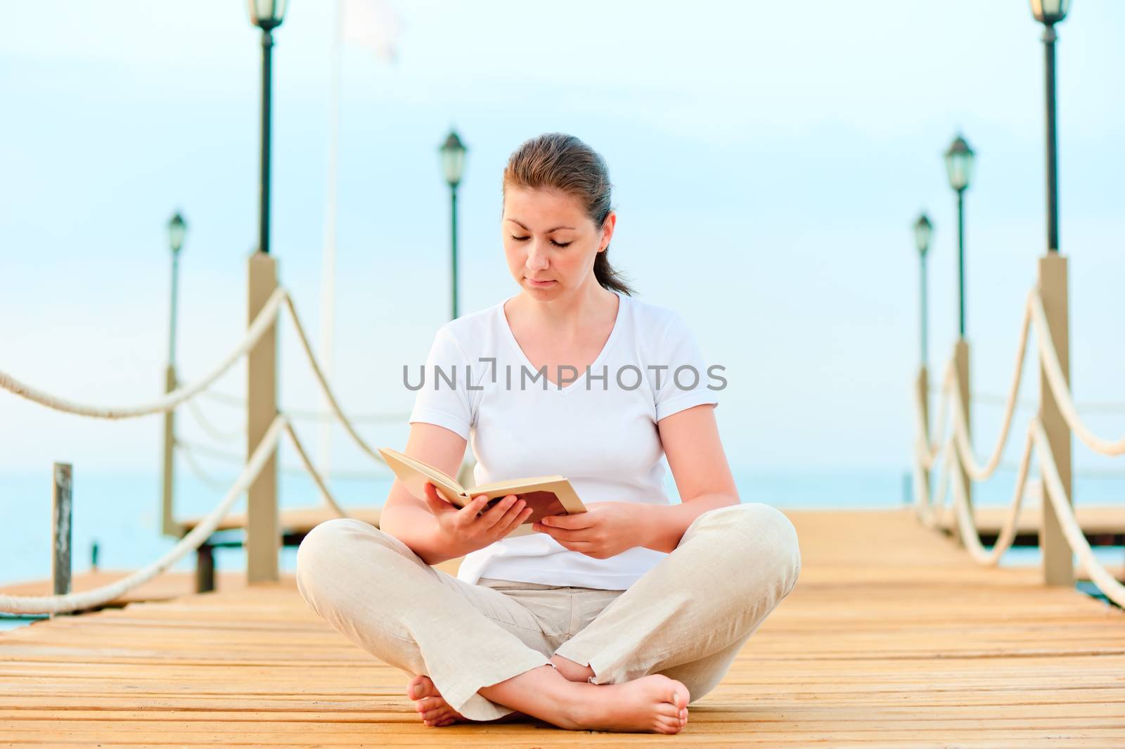 Young girl reading a book while sitting on a pier barefoot