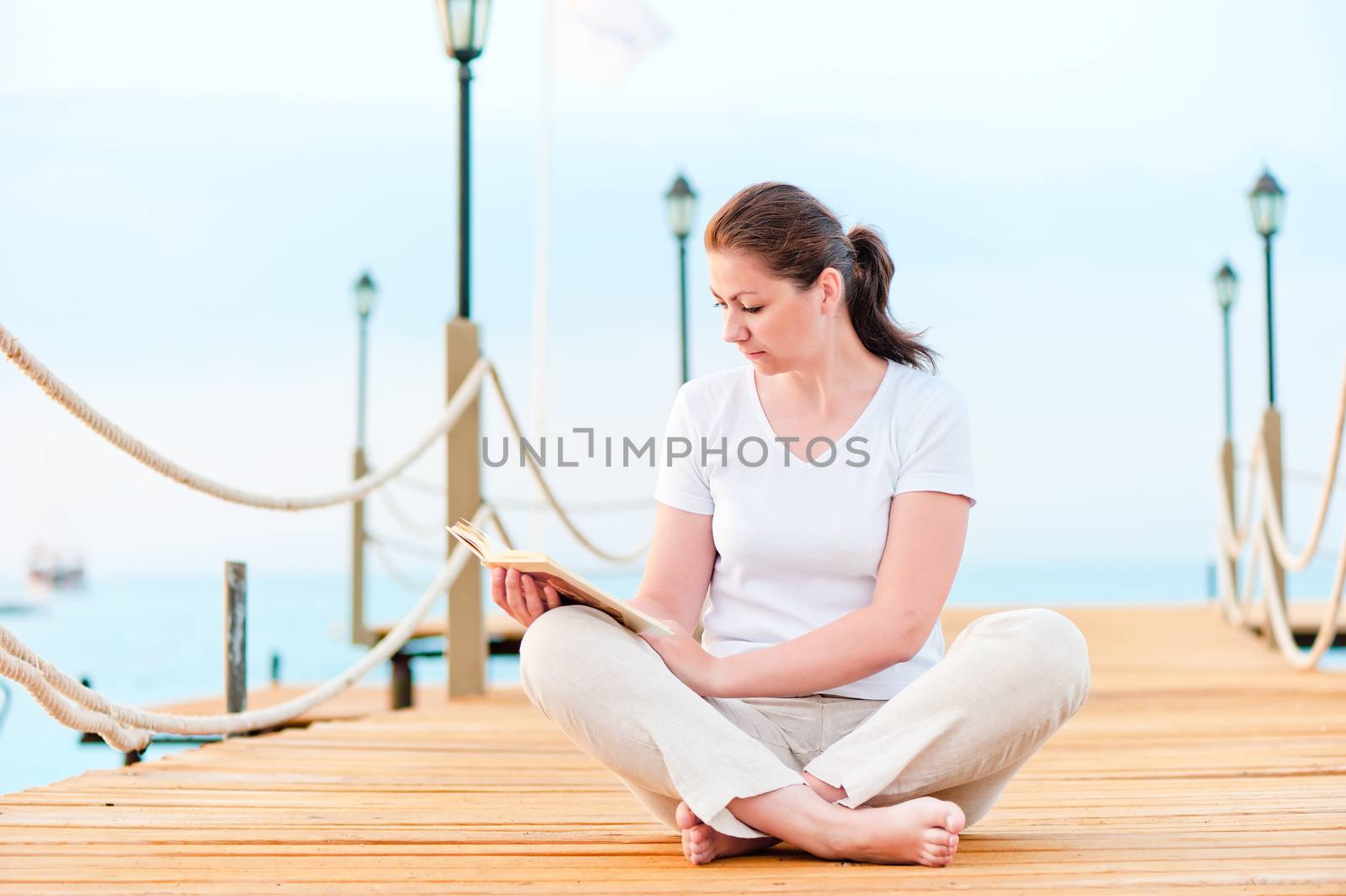 Young girl reading a book while sitting on a pier barefoot