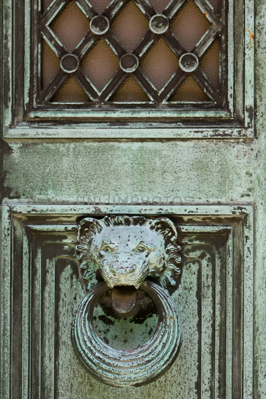 Close-up of a metal mausoleum door, weathered to a green patina, featuring a lion head doorknocker and frosted glass window.