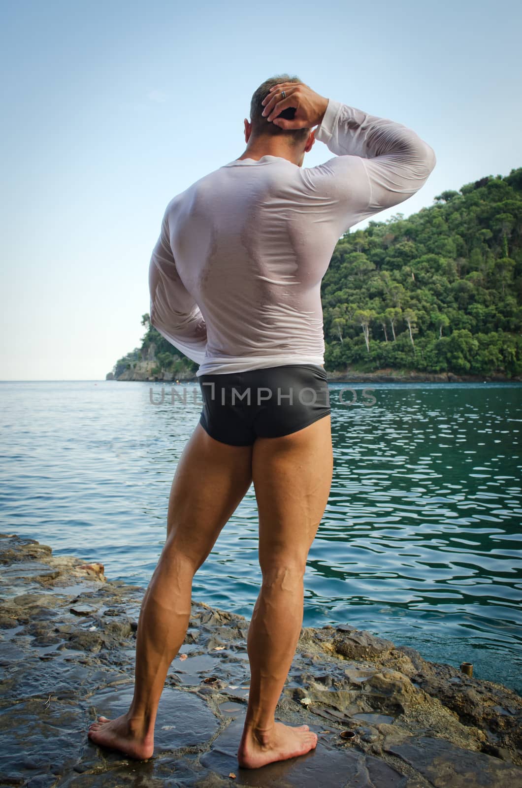 Muscular bodybuilder facing the sea, seen from the back wearing wet shirt