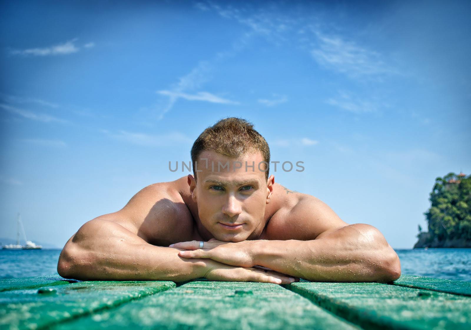 Muscular and handsome young man resting his chin on his hands, laying on wood planks looking at camera