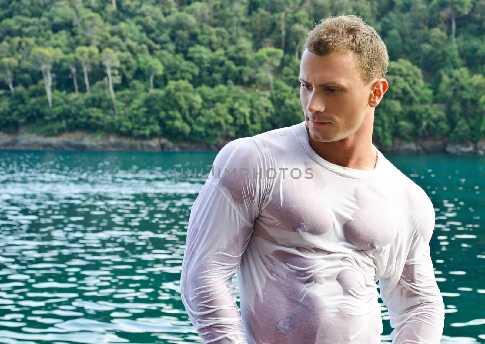 Attractive young bodybuilder by the sea with wet shirt on, serious expression