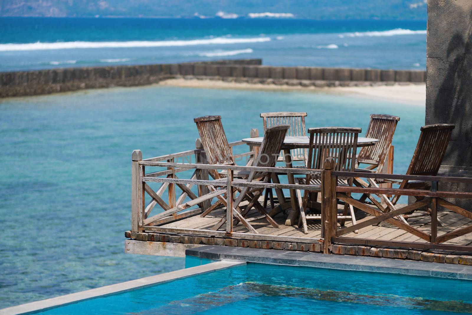 Wooden area with chairs and table near blue water of swimming pool and the sea on background
