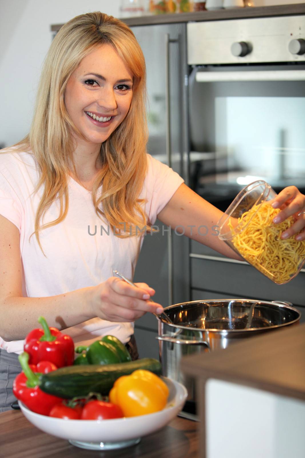Smiling pretty young blond housewife preparing pasta in the kitchen tipping noodles into a saucepan of boiling water on the stove
