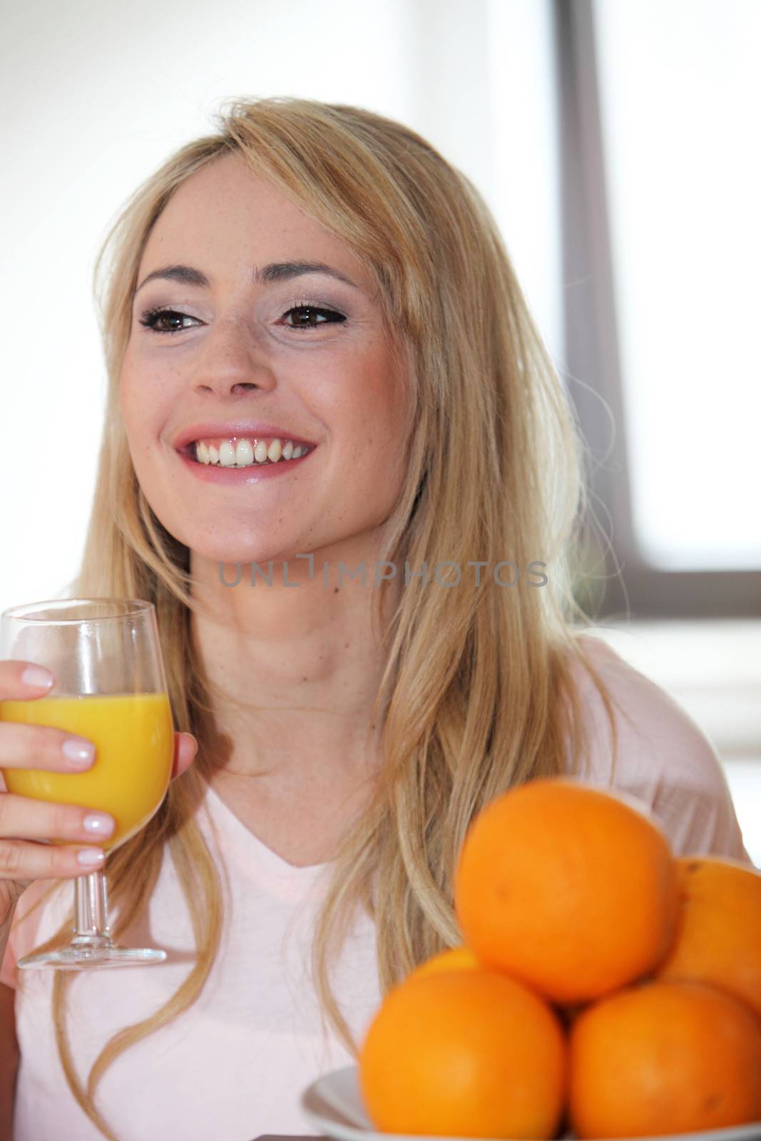 Smiling happy young woman enjoying a glass of fresh orange juice with whole oranges in a bowl in the foreground