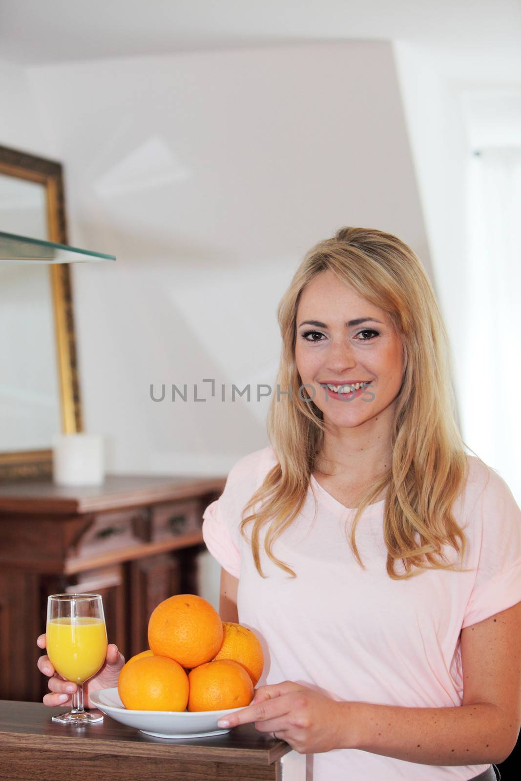 Smiling healthy young woman with a bowl of fresh oranges and a glass of freshly squeezed juice in her hand standing in her house