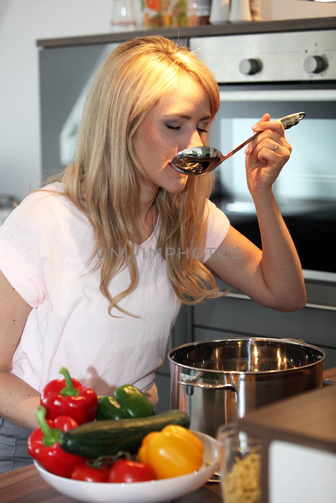 Pretty young blond woman standing over a saucepan on the stove tasting her cooking with a colourful bowl of fresh peppers in the foreground