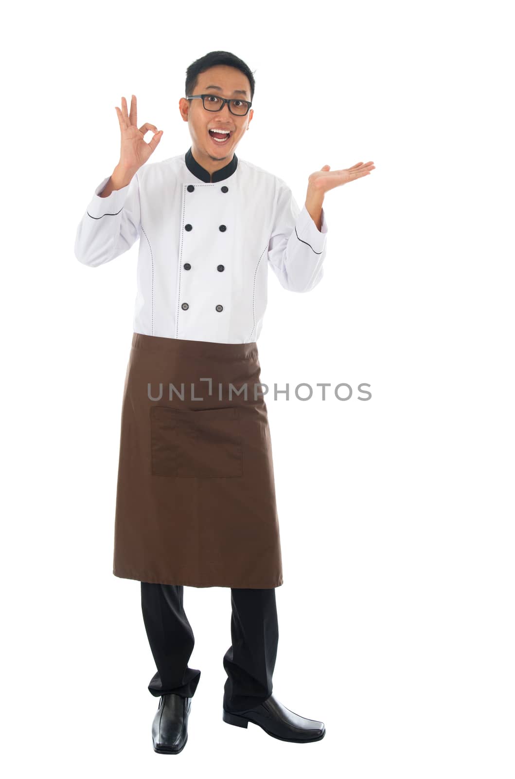 Portrait of full body Asian male chef showing copy space and okay hand sign, standing isolated on white background