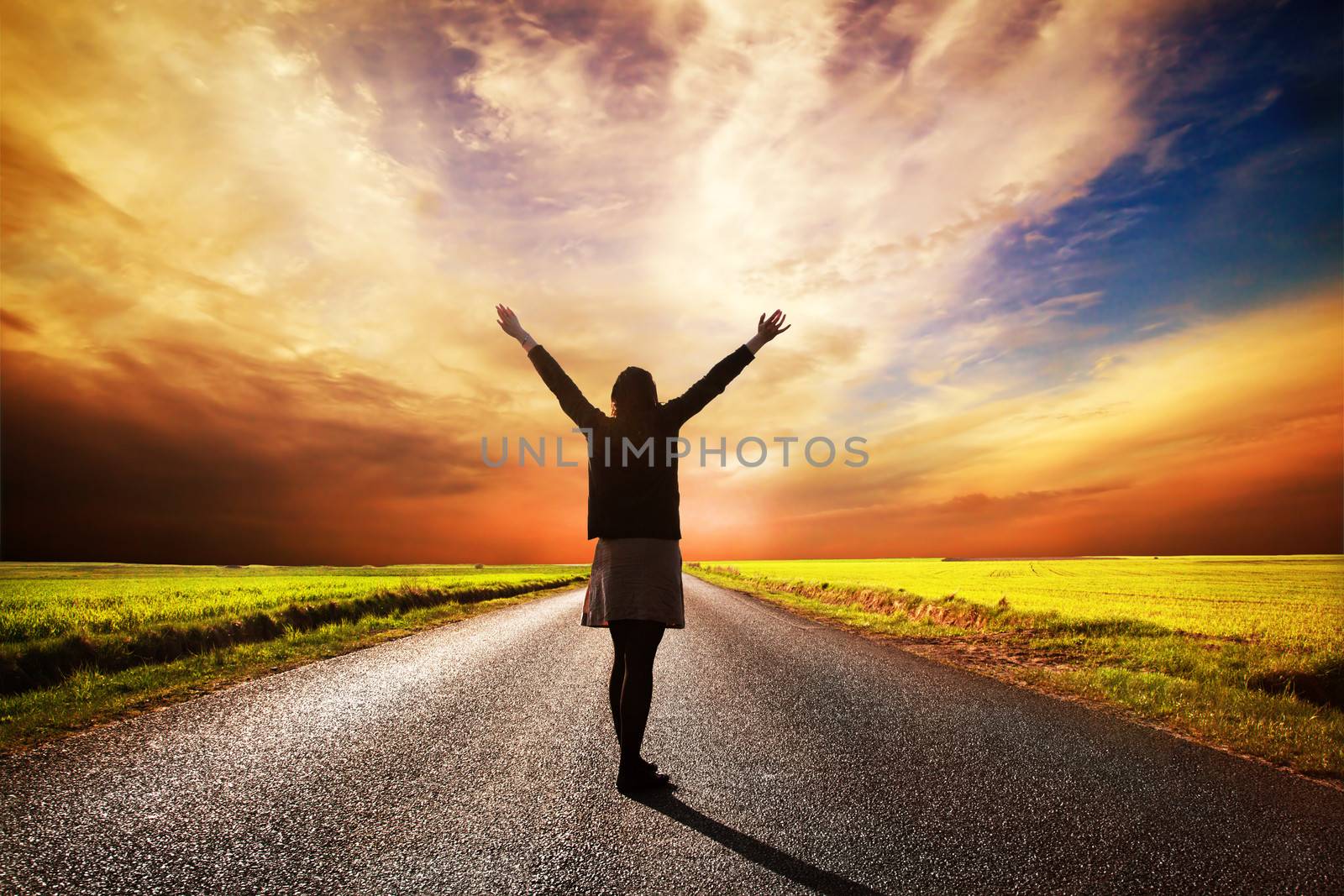 Happy woman standing with hands up on long straight road facing the sun. Sunset sky