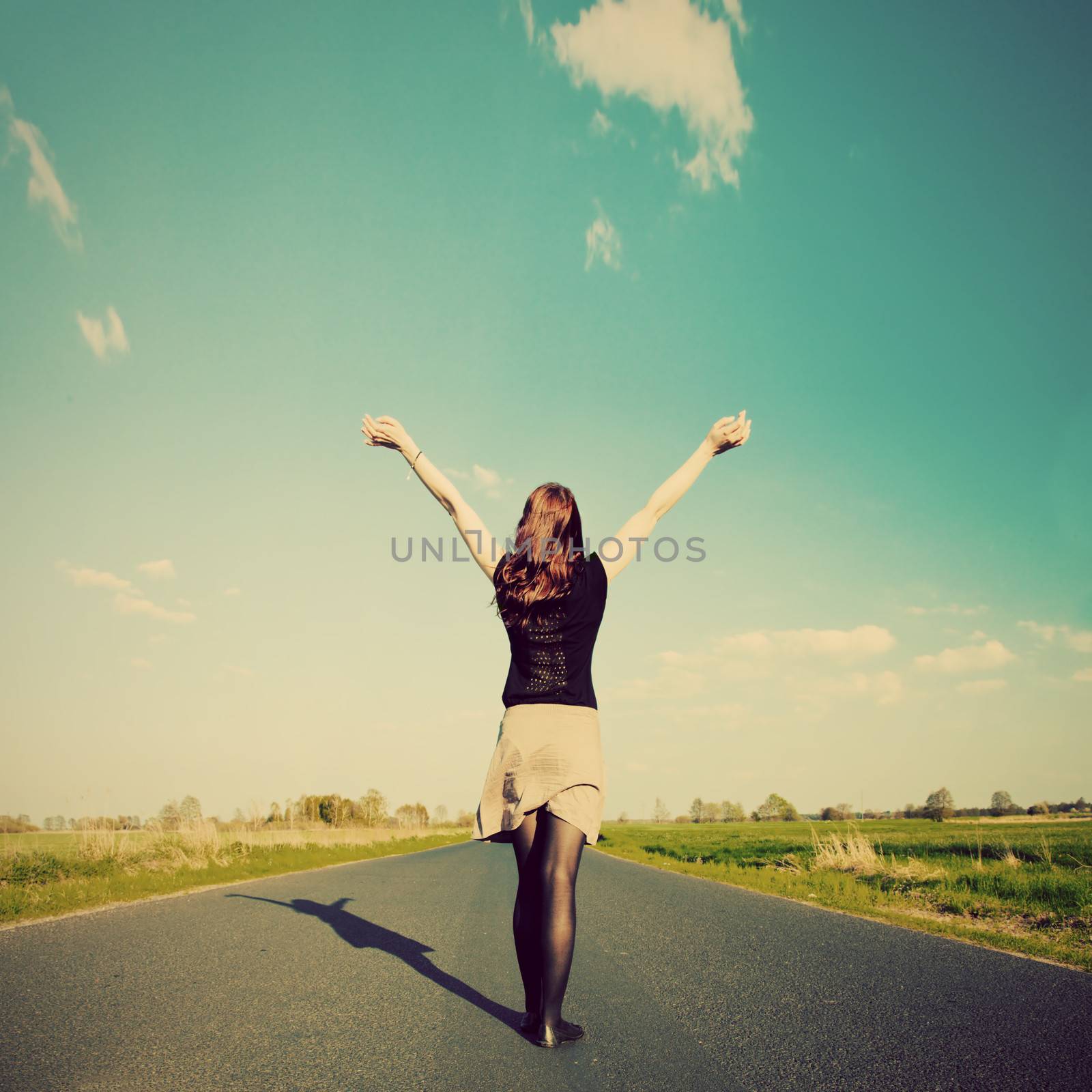 Happy woman standing with hands up on long straight road facing the sun. Retro vintage style