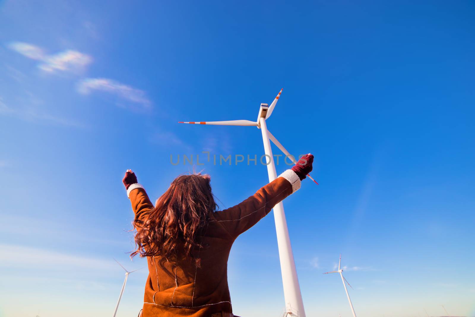 Young happy woman with hands up looking at wind turbines, eco energy source. Environment fiendly