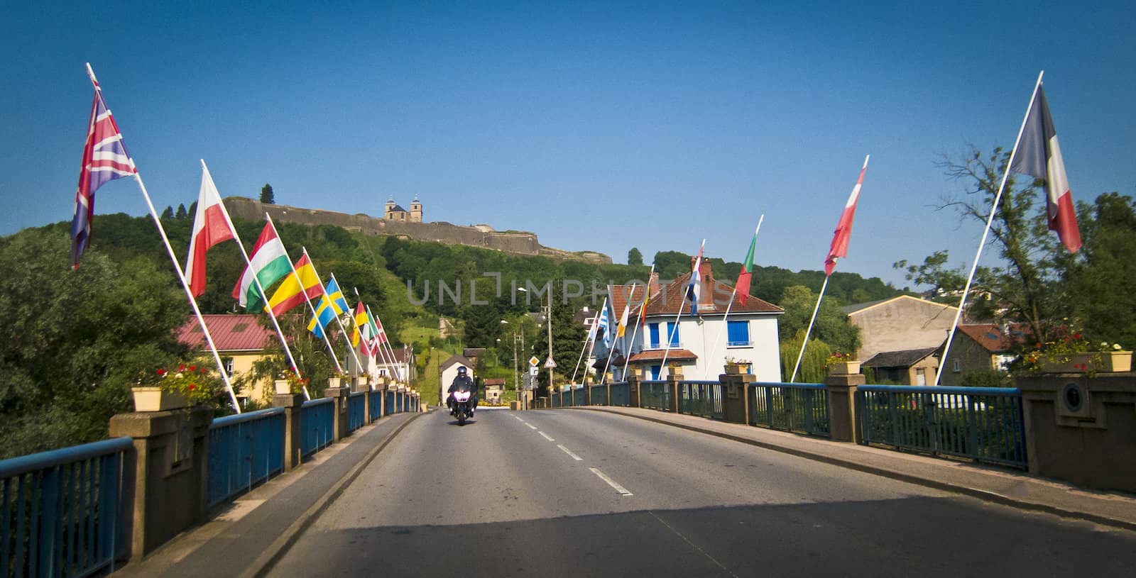 Bridge in France with european flags flying, a motorcyle approaching on bridge  and castle on the hill in the background