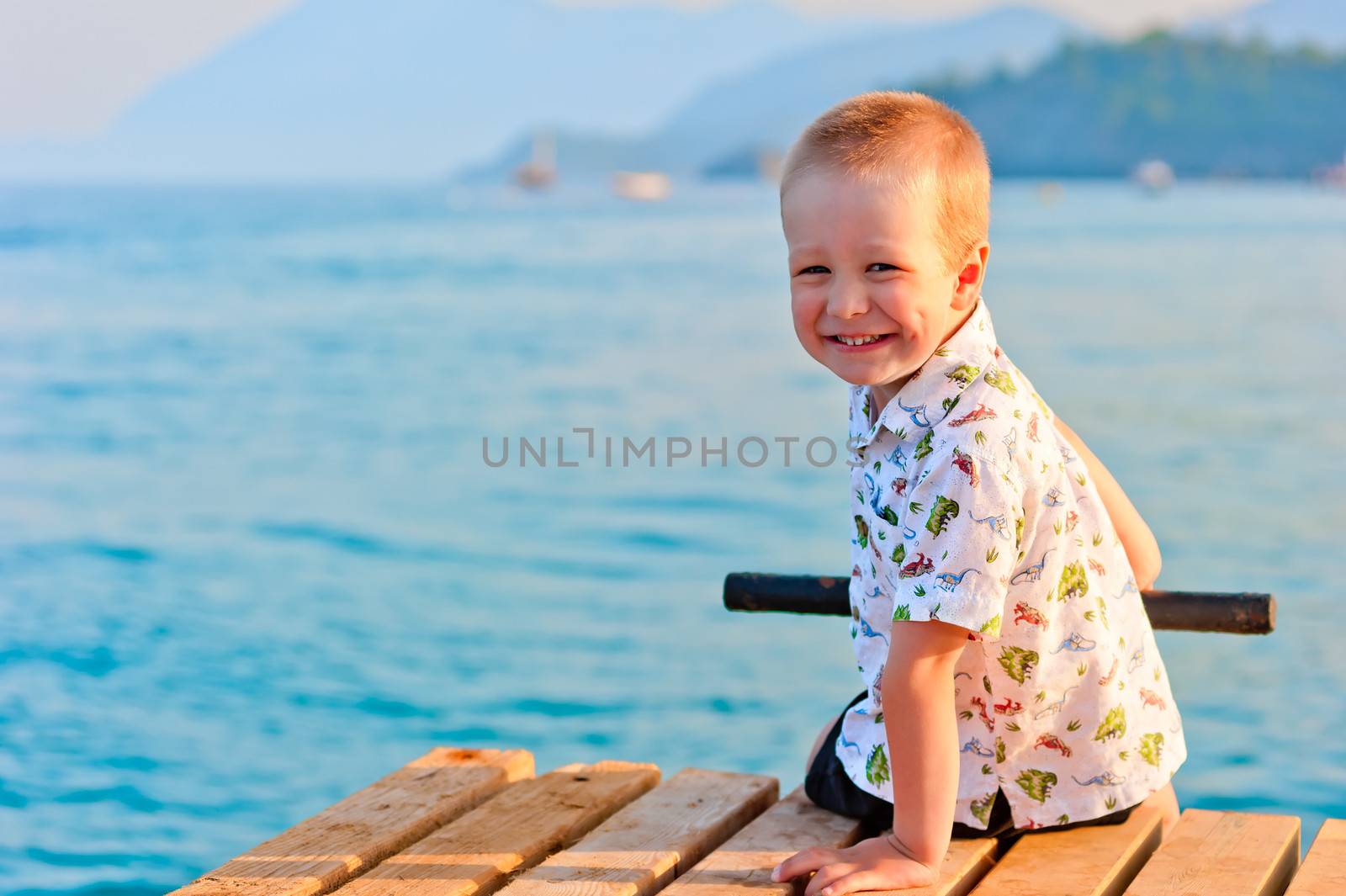 happy little boy sitting on a pier at sunset by the sea