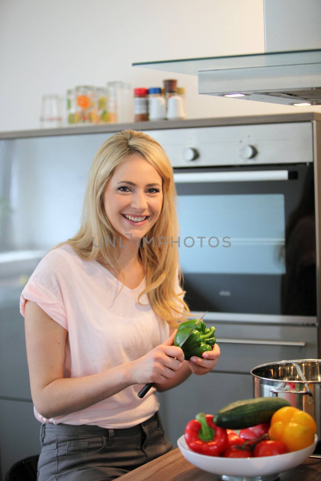 Happy woman cooking a meal in the kitchen cutting up a green pepper to put into a pot on the stove with a colourful bowl of fresh vegetables in the foreground