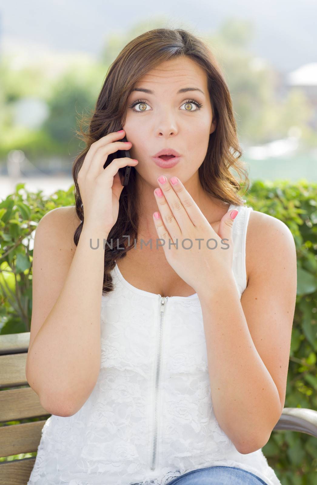 Shocked Young Adult Female Talking on Cell Phone Outdoors on Bench.