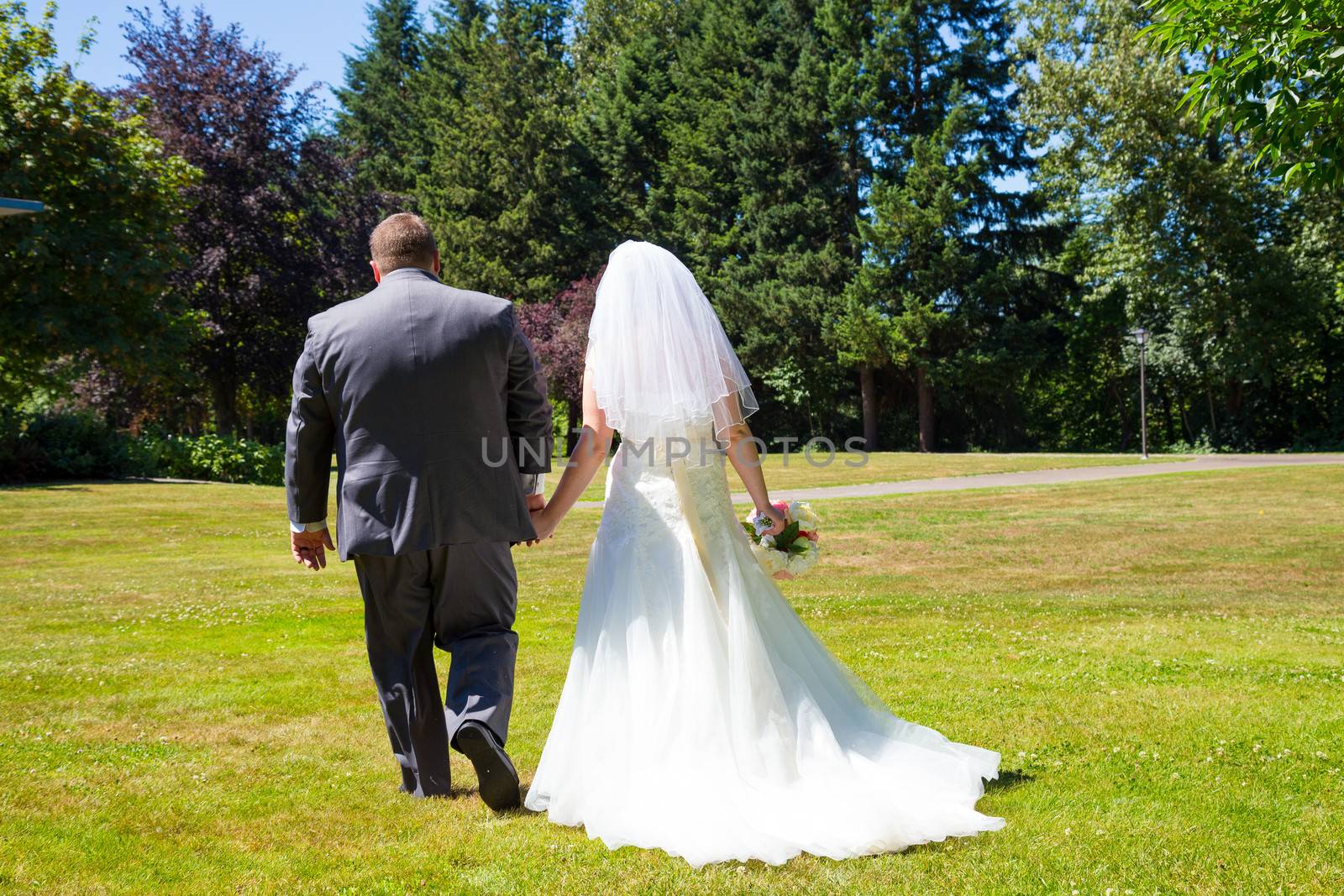 A bride and groom walk away from the camera holding hands at this outdoor wedding venue location in a park after just tying the knot.