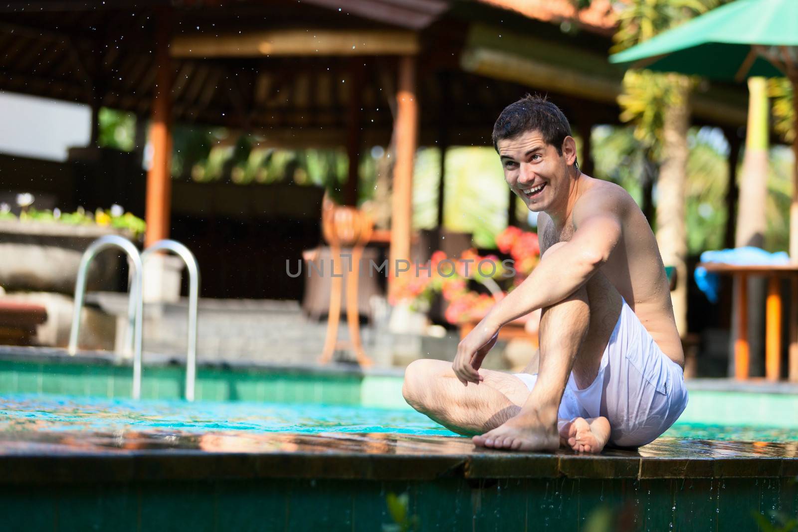 Happy young man sitting near swimming pool in tropical resort