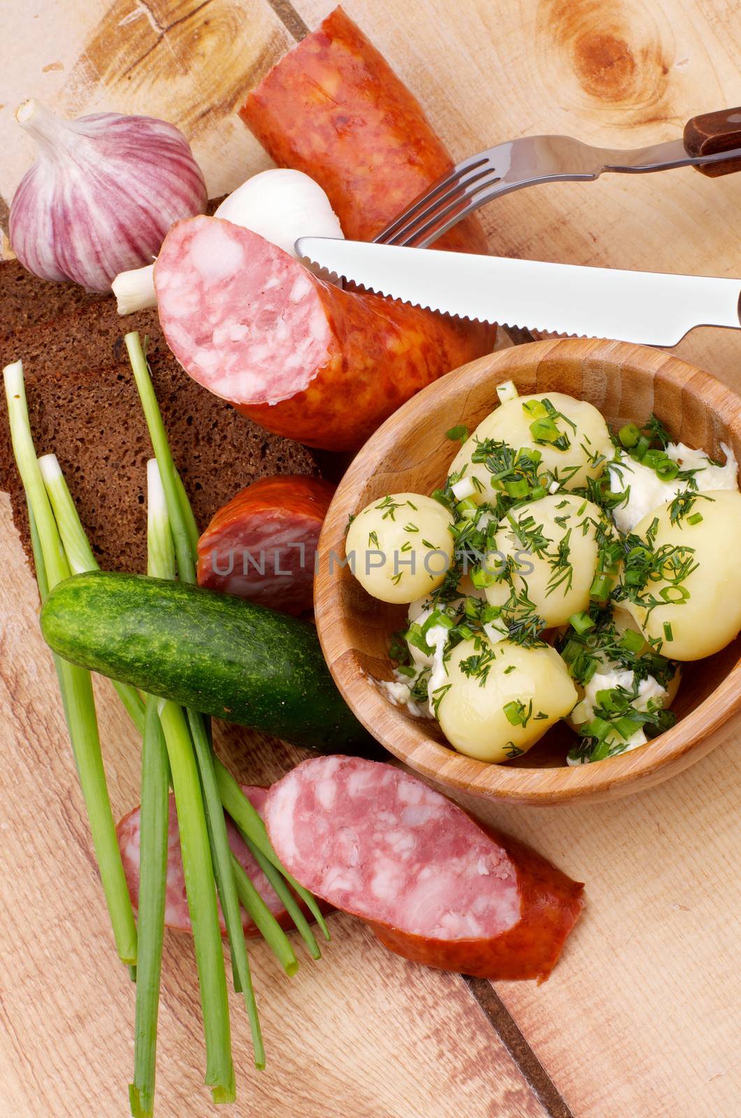 Arrangement of Boiled Potato, Spring Onion, Cucumber, Garlic, Homemade Smoked Sausage and Whole Grain Bread with Fork and Knife on Wooden background. Top View