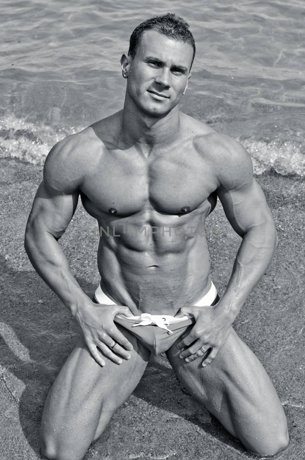 Handsome young bodybuilder in bathing suit kneeling on the beach by artofphoto