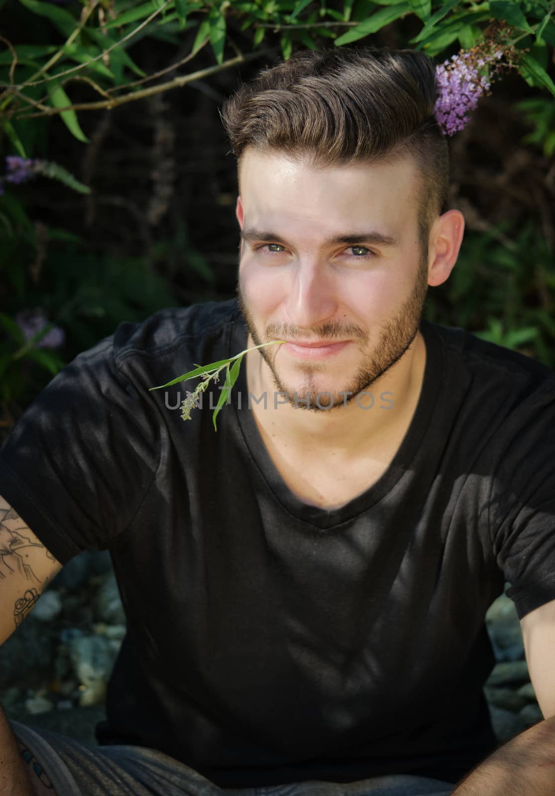 Attractive, smiling young man outdoors with flower between his lips, looking in camera